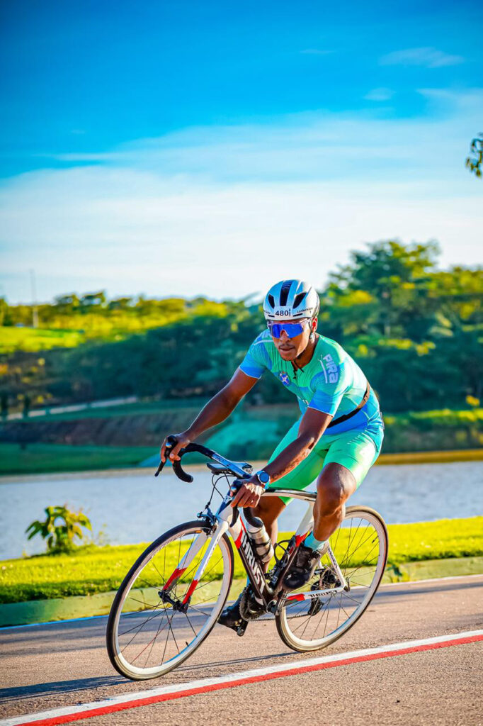 O ciclista Pedro Azevedo dos Santos pedala numa bicicleta de estrada durante a sua participação na prova de duatlo, usando capacete, óculos de sol e equipamento desportivo verde e azul.