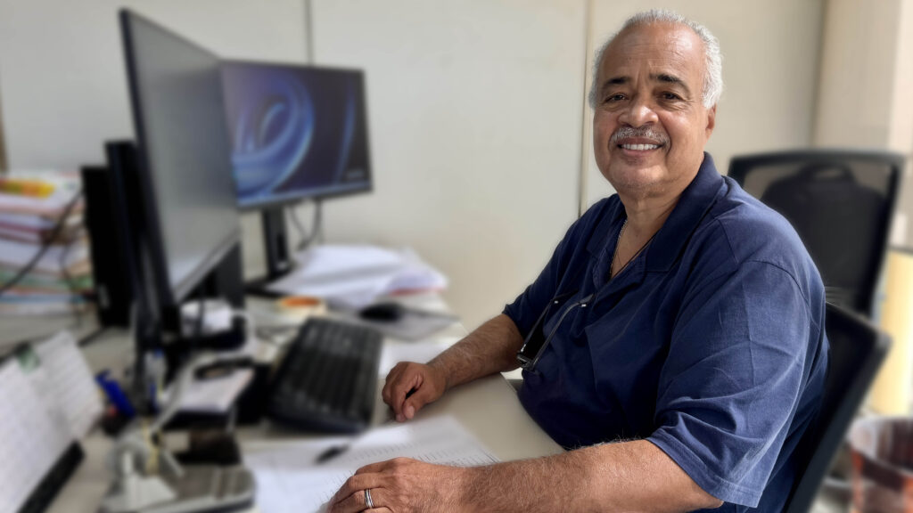 A imagem mostra Pedro Luiz Fernandes, assessor da Secretaria de Finanças da Prefeitura de Piracicaba, sentado a uma secretária de trabalho num escritório. Ele está a sorrir para a câmara, vestindo uma t-shirt polo azul-escura, num ambiente de trabalho equipado com monitores de computador, teclado e documentos.