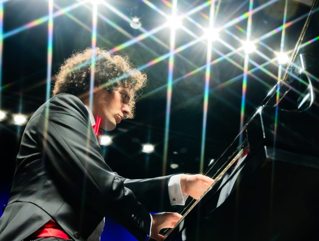 O pianista Fabio Martino é visto de perfil, de fato escuro e gravata borboleta vermelha, a tocar expressivamente um piano de cauda preto num palco iluminado por holofotes com efeito de brilho em estrela (starburst).
