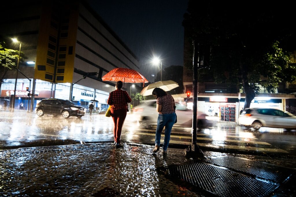 Cena urbana durante uma forte chuva noturna. Duas pessoas atravessam uma rua com guarda-chuvas em uma faixa de pedestres, enquanto carros passam ao fundo. As luzes dos postes, dos veículos e dos estabelecimentos refletem no asfalto molhado, destacando o volume da chuva.