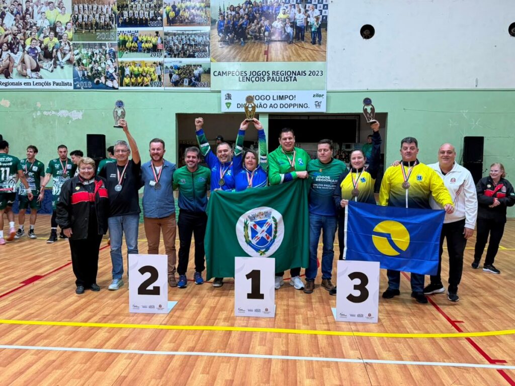 Atletas e dirigentes de várias comitivas posam num pódio numerado (1º, 2º e 3º lugares) num pavilhão desportivo, com a equipa vencedora ao centro a erguer a bandeira verde de Piracicaba e um troféu.