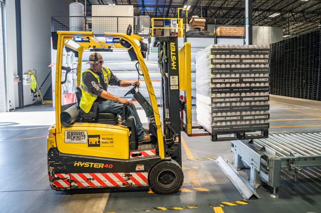 Mostra um operador com capacete e colete de segurança a manobrar uma empilhadeira amarela (marca Hyster) no interior de um armazém industrial. O veículo está a elevar e posicionar uma palete carregada com caixas de mercadoria sobre uma esteira rolante de transporte.