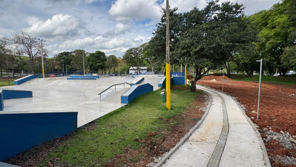 Vista de nível do solo mostrando a pista de skate recém-reformada com estruturas azuis e, ao lado, a nova pista de caminhada em betão com piso tátil a ser construída contornando o espaço.
