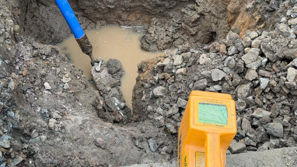 Apresenta um detalhe em plano aproximado da técnica de furo direcional subterranean, mostrando a tubulação azul a ser puxada pelo solo e um equipamento localizador amarelo em primeiro plano.