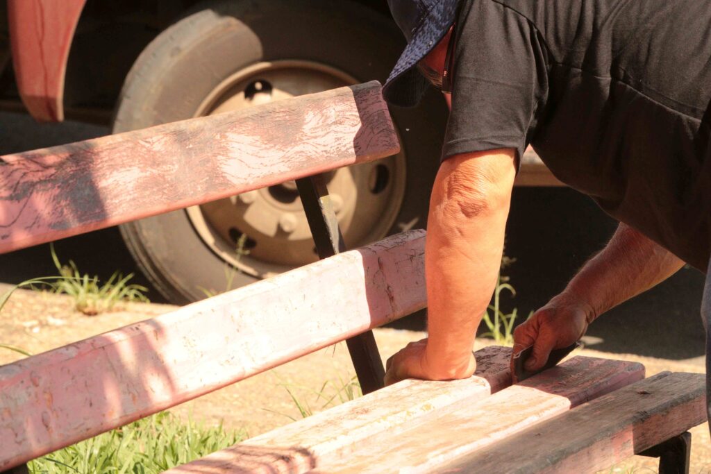 Um trabalhador aparece lixando o encosto e o assento de madeira de um banco público para restauração e pintura.