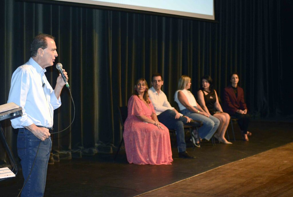 O prefeito Helinho Zanatta a discursar com um microfone na lateral do palco, enquanto os demais membros da mesa de honra acompanham sentados ao fundo.