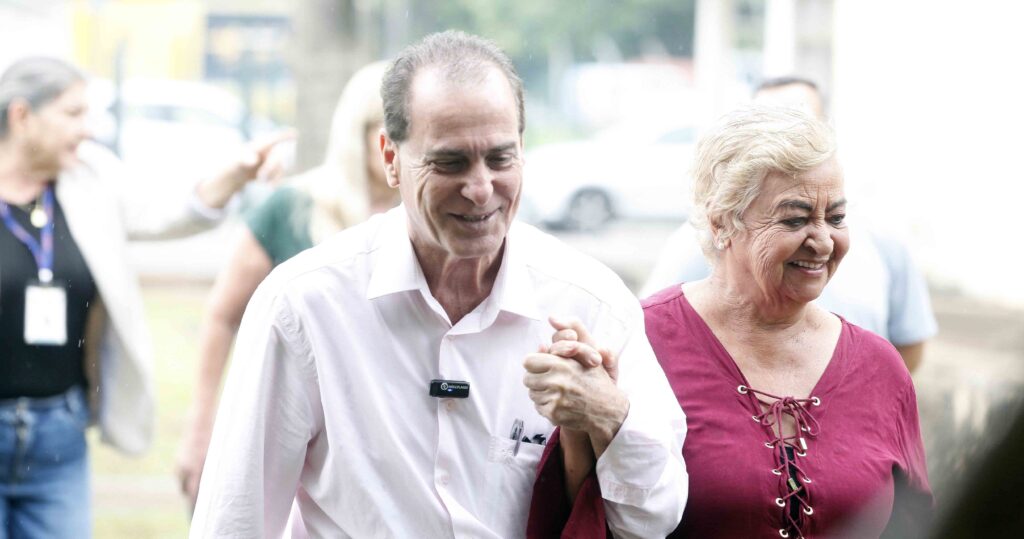 Um homem e uma mulher caminham sorridentes de mãos dadas num evento ao ar livre.