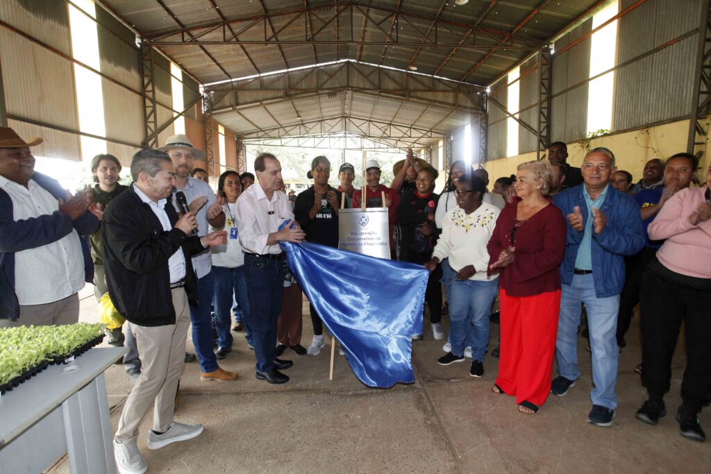 Momento de inauguração num galpão, onde um grupo de pessoas aplaude enquanto uma placa comemorativa é desvelada ao retirar-se um pano azul.