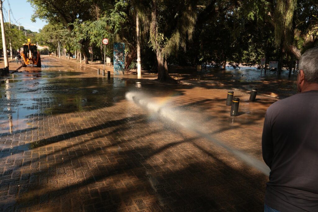 Exibe um plano mais amplo de uma avenida pavimentada com paralelepípedos, onde um jato potente de água está a lavar o piso, criando um pequeno arco-íris com a luz do sol. No lado esquerdo, avista-se uma retroescavadeira ao fundo, enquanto a vegetação que margeia o rio surge do lado direito.
