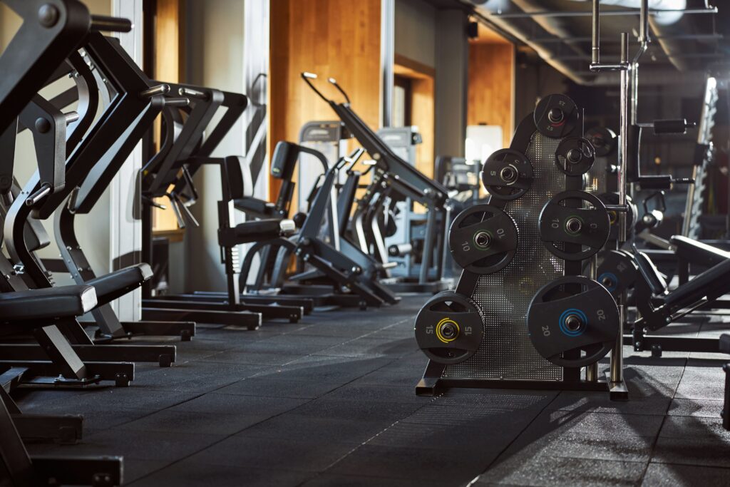 Interior de uma academia de musculação moderna, destacando em primeiro plano um suporte vertical com anilhas e pesos pretos organizados por tamanho, ao lado de diversos aparelhos e equipamentos de treino dispostos sobre o piso emborrachado.