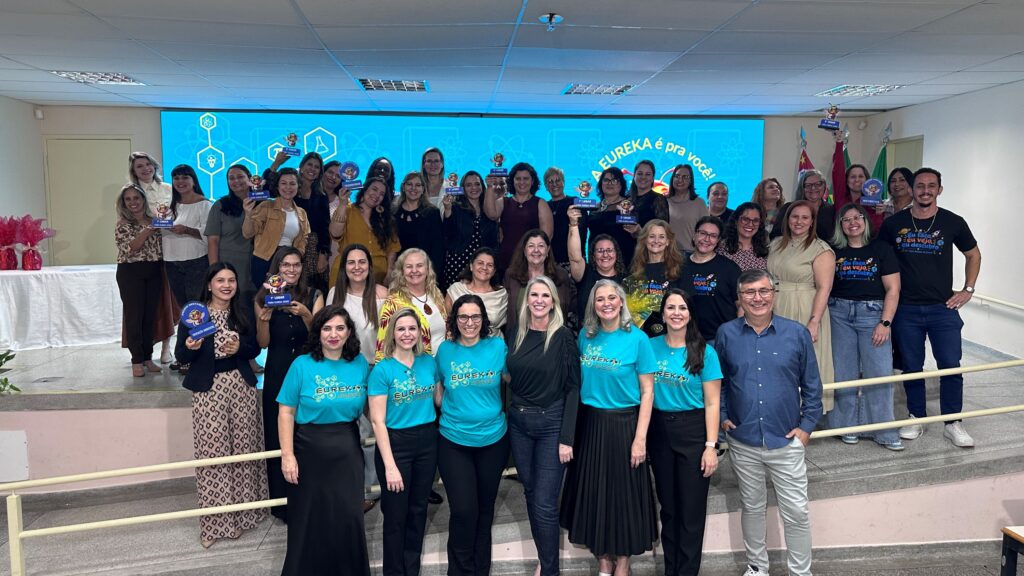 Uma fotografia de grupo ampla que reúne os professores premiados e organizadores da Feira Eureka, posando juntos num auditório com troféus nas mãos.
