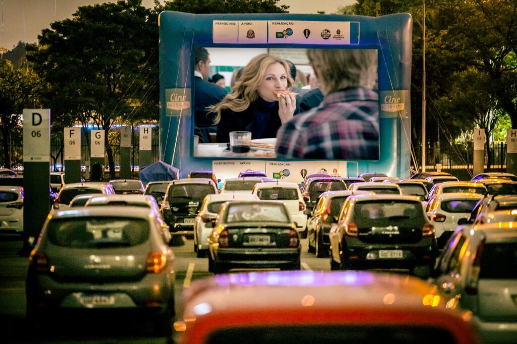 Plano aproximado de um ecrã de cinema insuflável que projeta uma cena do filme Comer, Rezar, Amar (com Julia Roberts). Vários carros estão estacionados em filas organizadas no recinto a assistir à exibição.