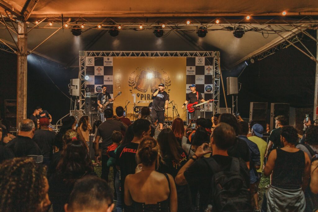 Uma vista do público a assistir a um concerto noturno sob uma tenda decorada com luzes suspensas. No palco, uma banda composta por vocalista e músicos (guitarrista e baixista) atua em frente a um painel do "Festival de Cerveja".