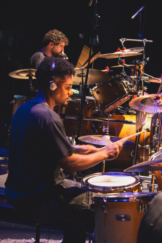 Um homem adulto com auscultadores toca bateria num palco iluminado, enquanto outro baterista aparece ao fundo, também a tocar.