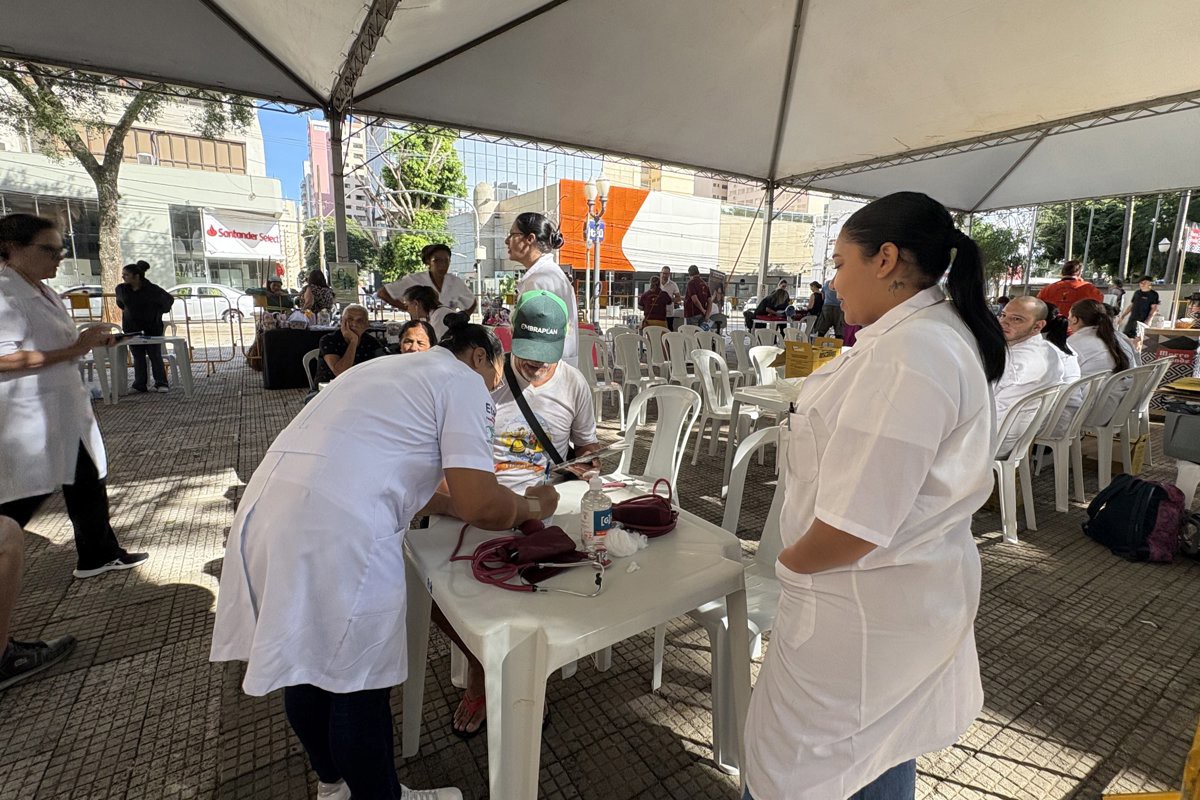 Apresenta outro ângulo de um evento de atendimento à comunidade sob uma grande tenda num espaço público. Em destaque no primeiro plano, uma profissional de saúde de bata branca atende um homem sentado à mesa, enquanto outra profissional acompanha o atendimento em pé. Ao redor, há diversas cadeiras de plástico brancas dispostas e outros atendimentos a decorrer.