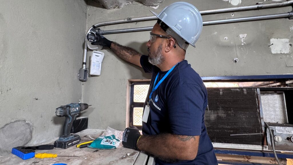 Um trabalhador com capacete de segurança branco, óculos de proteção e blusa azul ajusta uma câmara de segurança instalada na parede junto a calhas elétricas, tendo à sua frente uma bancada com ferramentas e um berbequim.