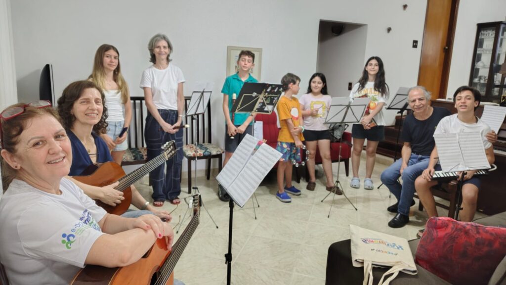 Grupo de alunos e professores reunidos numa sala com instrumentos musicais, como violões, flautas doces e estantes de partitura, para um ensaio de recital.