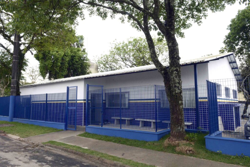 A fachada exterior de um edifício público de saúde pintado de branco e azul, protegido por grades metálicas azuis e cercado por árvores.