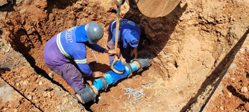 Dois trabalhadores com equipamento de proteção individual encontram-se dentro de uma vala a manusear e alinhar um conjunto de tubagens e uma nova válvula azul. A peça está a ser içada por uma cinta suspensa por uma máquina externa.