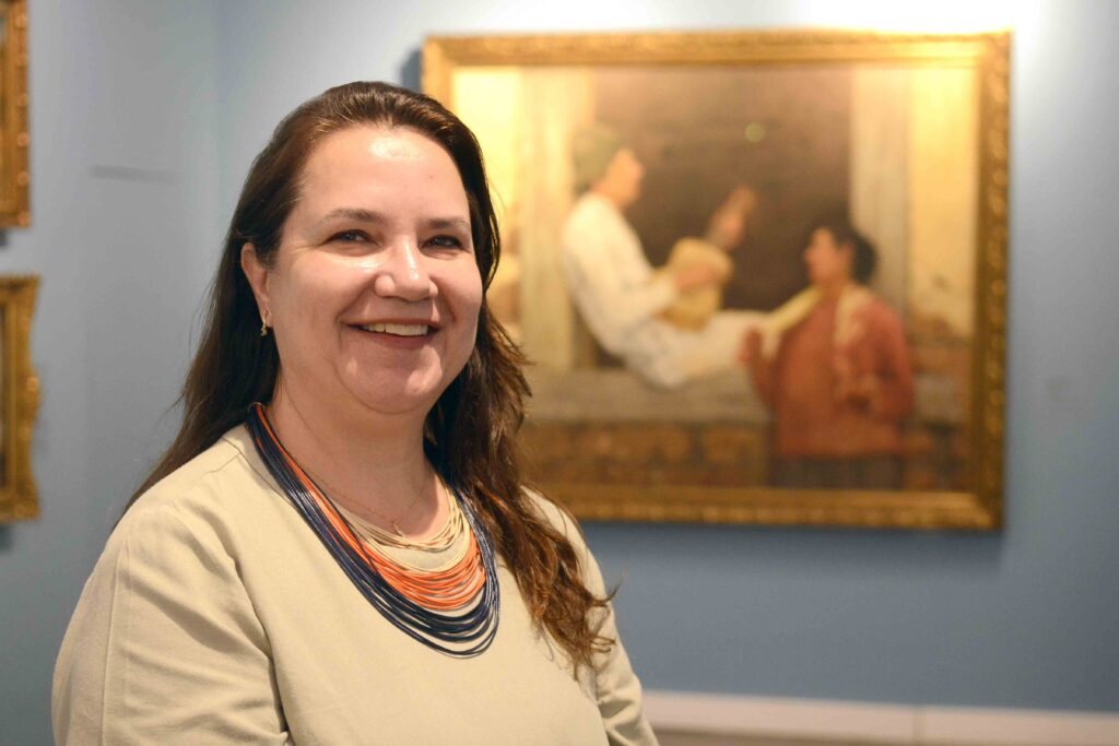 A responsável pela Pinacoteca Miguel Dutra, Renata Gava, a sorrir em primeiro plano, tendo ao fundo a famosa pintura O Violeiro numa parede azul.