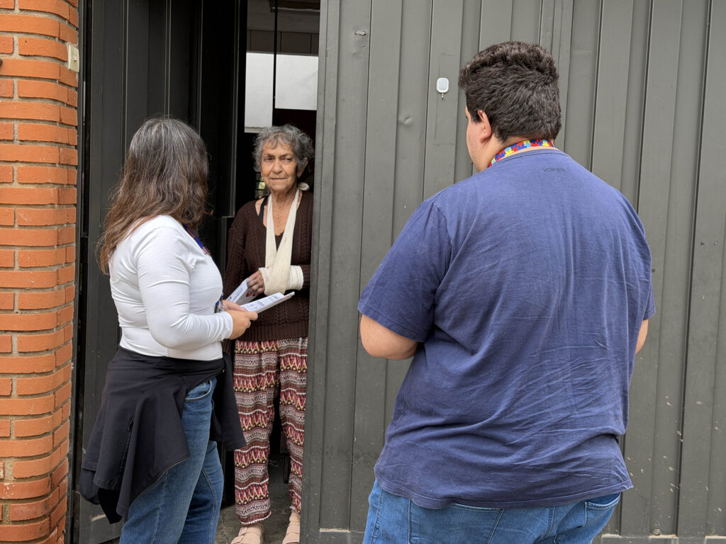 Dois agentes de orientação conversam ao portão de uma residência com a moradora Márcia Rocha, entregando folhetos educativos.