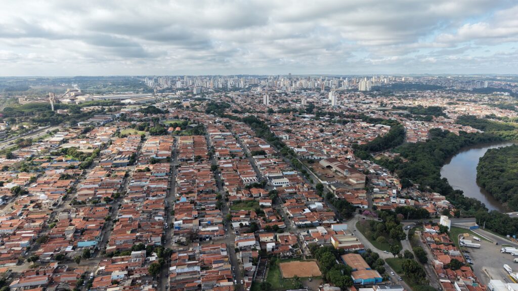 Vista aérea panorâmica da cidade de Piracicaba, destacando a malha urbana com as suas casas, prédios ao fundo e o curso do rio a rodear a vegetação local.
