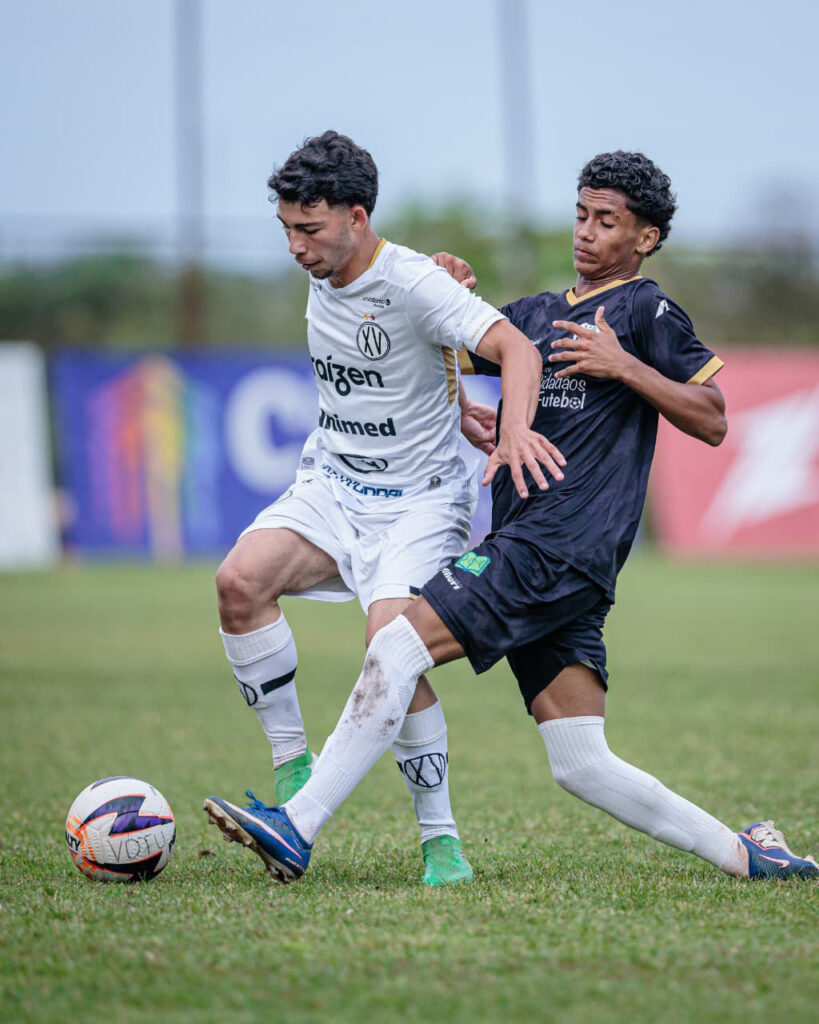 Jogador de futebol da equipa sub-20 do XV de Piracicaba, de uniforme branco, disputa a bola no relvado com um adversário de uniforme escuro.
