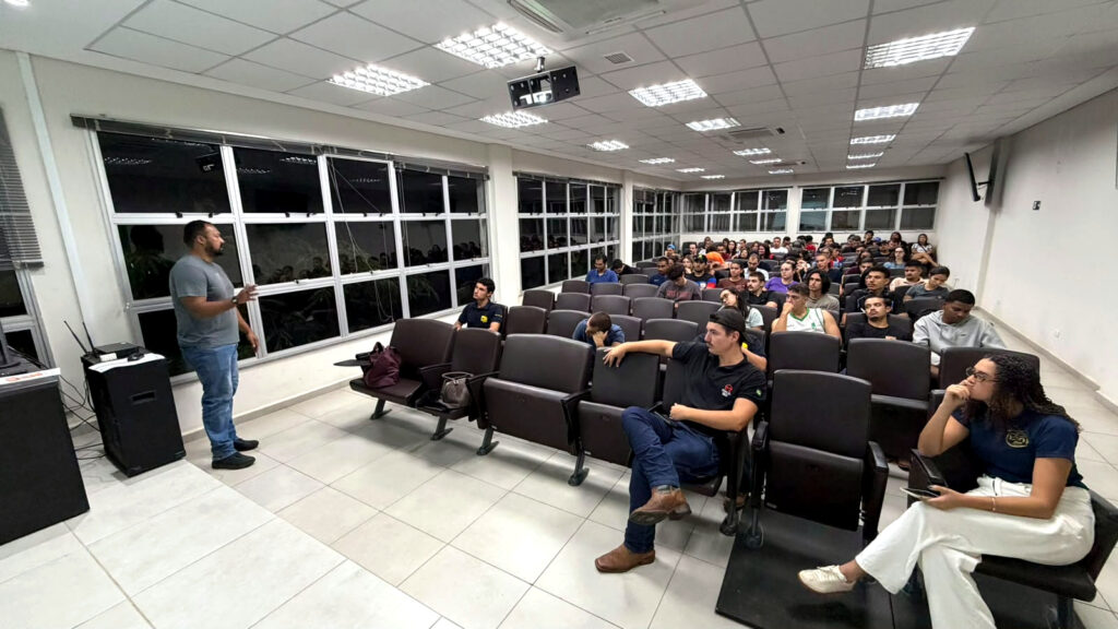 Um homem está de pé a falar para uma plateia num auditório bem iluminado, preenchido com várias fileiras de cadeiras escuras onde se encontram sentados vários estudantes.