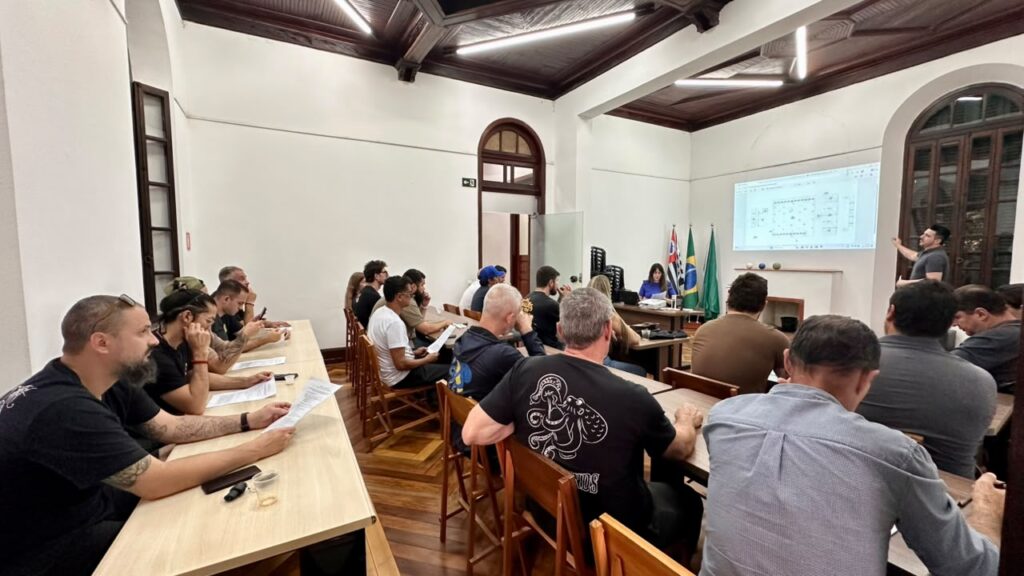 Uma reunião institucional numa sala ampla com mesas de madeira organizadas em filas, onde vários participantes acompanham uma apresentação projetada na parede ao fundo.