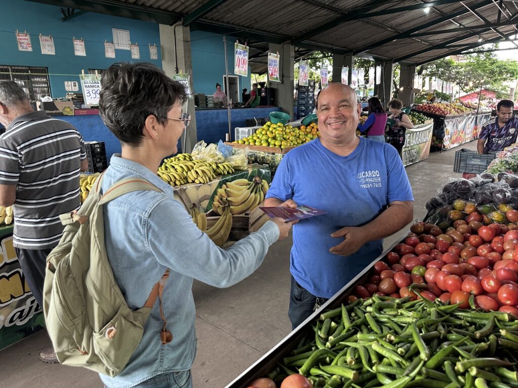 Uma agente entrega um folheto informativo ao feirante Ricardo Rodrigues Ribeiro, que sorri atrás da sua banca de vegetais e frutas num mercado municipal/varejão.