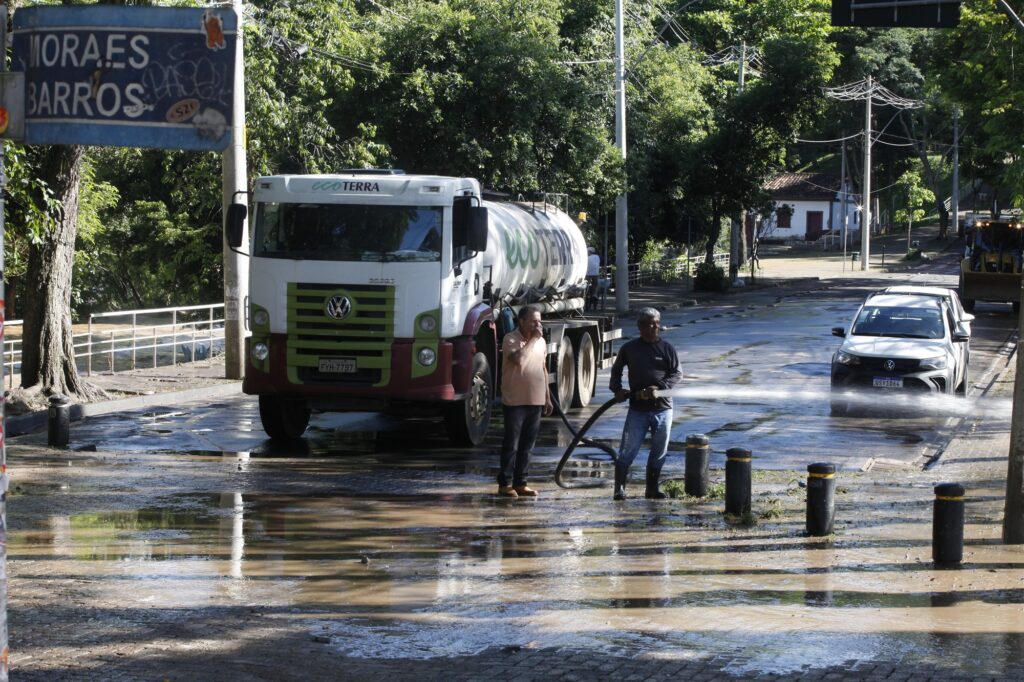 Apresenta uma rua coberta por lama e poças de água, com um caminhão-cisterna (EcoTerra) parado. Ao lado do veículo, dois trabalhadores utilizam uma mangueira de alta pressão para lavar a via. Em segundo plano, vê-se um carro branco a circular e vegetação densa ao fundo.