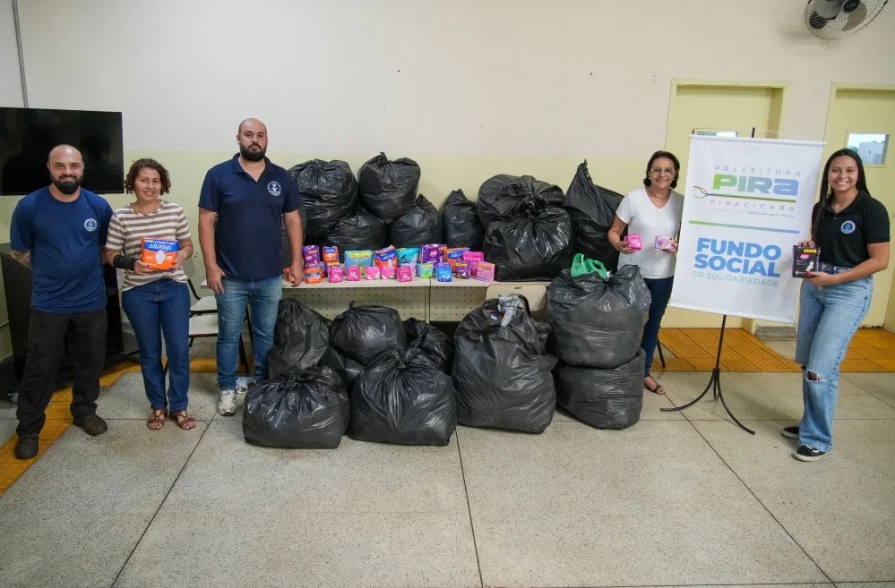 Cinco pessoas estão posadas lado a lado num espaço interior, ao pé de um painel informativo do Fundo Social de Solidariedade da Prefeitura de Piracicaba. No chão e sobre as mesas ao fundo, veem-se dezenas de sacos pretos grandes e embalagens de produtos de higiene feminina (pensos higiénicos e fraldas) doados. Alguns dos presentes seguram embalagens destes produtos enquanto sorriem para a câmara.