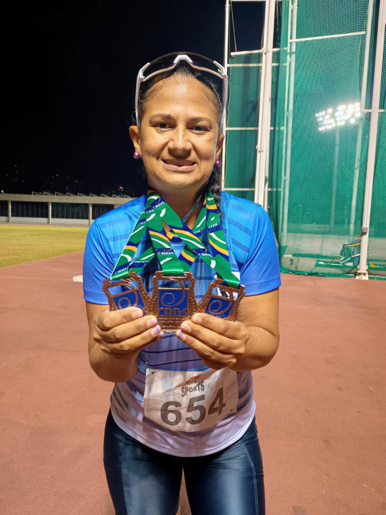 A atleta Rosângela Alves Ramos surge sorridente numa pista de atletismo, utilizando a indumentária desportiva com o número de peito 654 e óculos de sol na cabeça, enquanto exibe orgulhosamente no peito três medalhas de bronze conquistadas na competição.