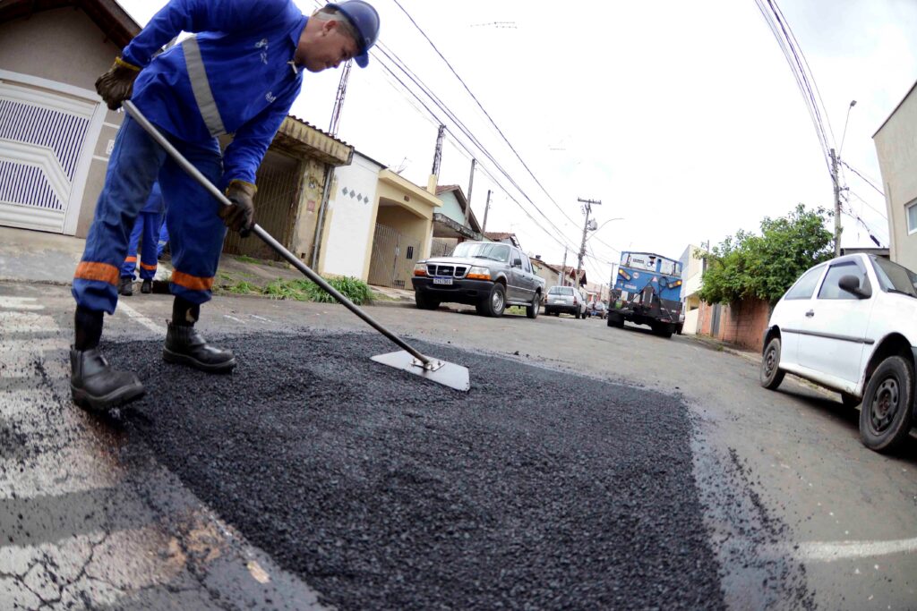 A imagem mostra um trabalhador de vestuário azul, capacete e luvas de proteção a utilizar uma ferramenta manual para nivelar e espalhar asfalto fresco numa secção da estrada. Ao longo da rua residencial, sob um ângulo de grande angular, sobressaem casas, um carro branco estacionado em primeiro plano, uma camionete e um camião de serviço mais ao fundo.