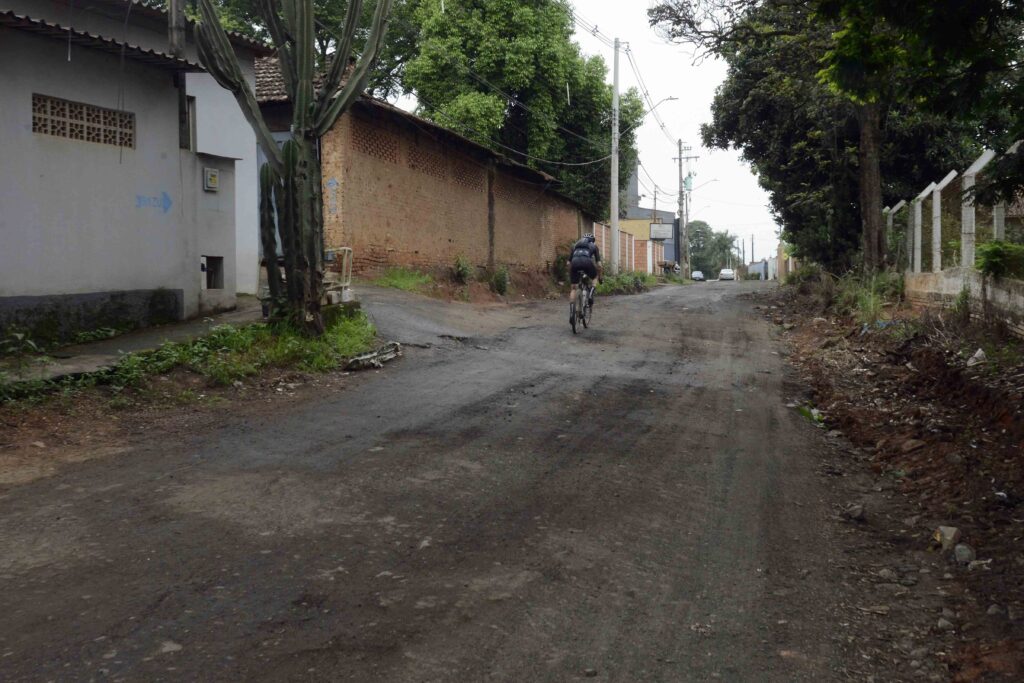 Rua de asfalto irregular e com terra batida, com um ciclista a circular ao centro ao lado de muros e vegetação.