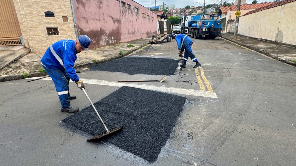 Trabalhadores de uniforme azul e capacete realizam o nivelamento e a reparação do asfalto numa rua do bairro Jaraguá, com um camião de apoio estacionado ao fundo.