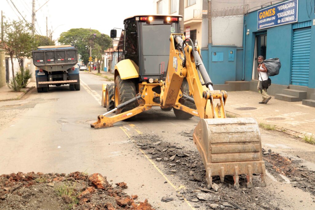 Uma retroescavadora amarela opera no meio de uma rua urbana asfaltada, realizando trabalhos de remoção e preparação do pavimento, com um camião estacionado ao fundo e um transeunte no passeio.