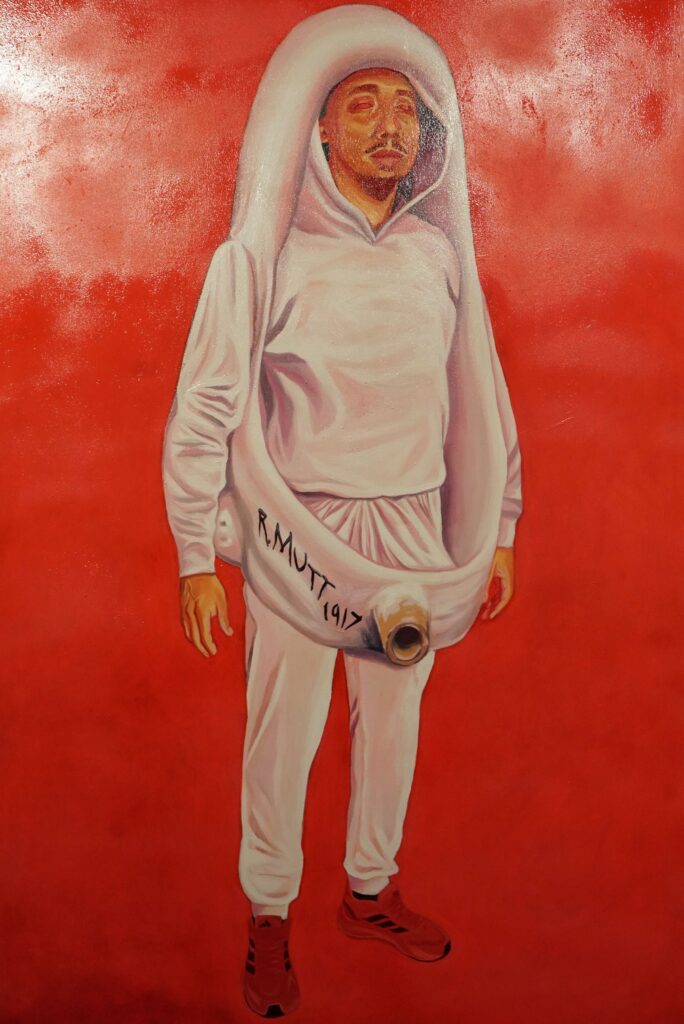 Apresenta uma pintura em fundo vermelho vivo com uma figura masculina no centro. O homem veste calças, camisola com capuz e sapatilhas de desporto claros, estando envolvido por uma estrutura branca curvada em formato de urinol que exibe a inscrição