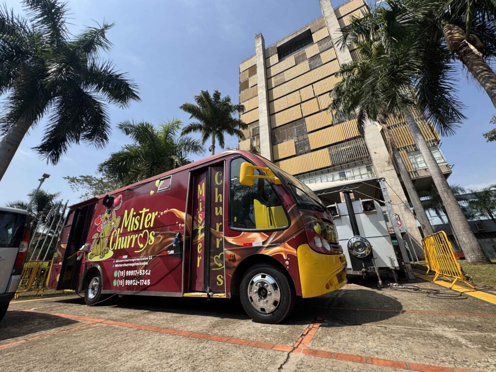 Um food truck de churros na cor vinho e amarelo estacionado em frente ao edifício do Centro Cívico (Prefeitura) de Piracicaba, cercado por palmeiras sob céu limpo