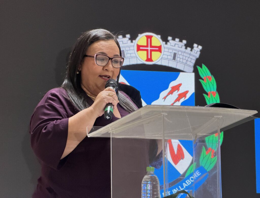 Uma educadora (Sandra Regina de Souza) discursa num púlpito transparente com microfone, em frente ao painel do evento com o brasão de Piracicaba ao fundo.
