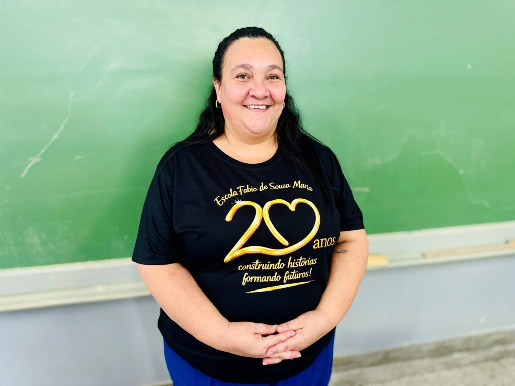 A educadora Sandra Rodrigues de Souza sorri enquanto faz um gesto de cumprimento (high five) com as mãos sobrepostas às de três estudantes numa mesa de biblioteca escolar.