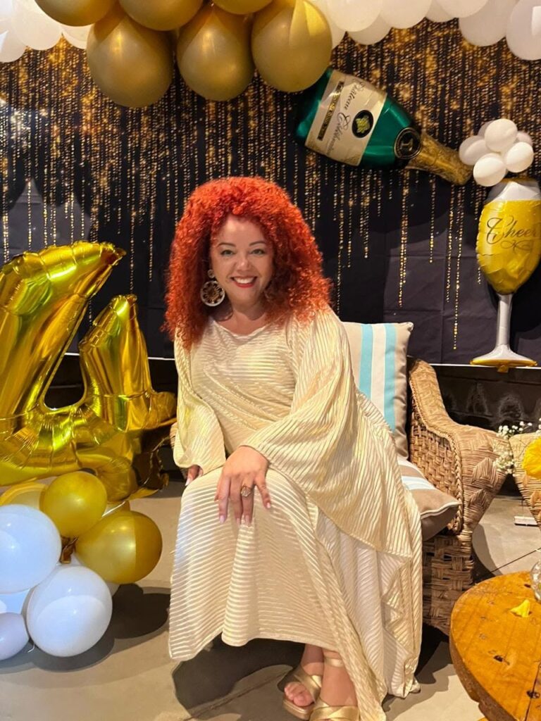 Uma mulher de cabelo encaracolado ruivo e vestido claro sorri sentada num espaço decorado com balões dourados e brancos e um painel brilhante ao fundo.