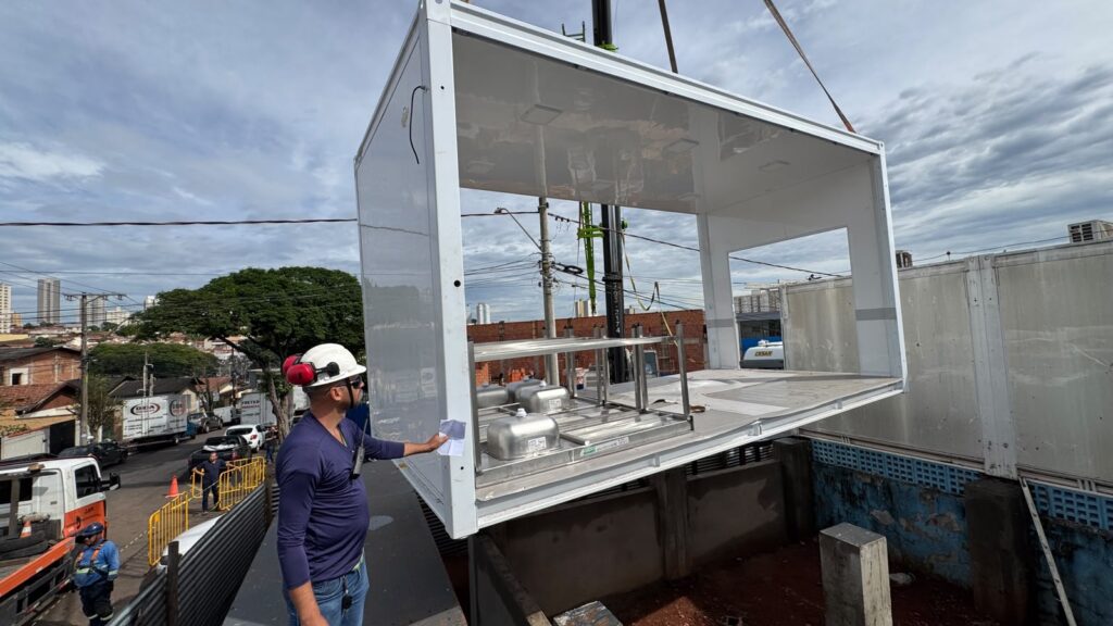 Um operador com capacete e abafadores de ruído orienta a manobra de um módulo pré-fabricado branco suspendo por grua. O módulo, equipado com bancadas e pias de aço inoxidável no interior, está a ser encaixado na estrutura do edifício.