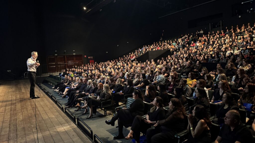 O psicoterapeuta e escritor Leo Fraiman ministra uma palestra sobre saúde mental no palco para uma grande plateia no auditório.