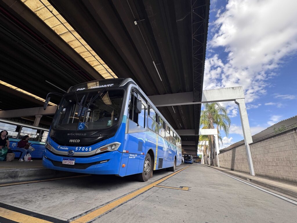 Um autocarro do transporte público de cor azul e cinzenta está estacionado numa plataforma coberta do terminal urbano, sob um céu ensolarado e poucas nuvens. Ao fundo, observam-se passageiros aguardando no terminal e palmeiras junto às instalações.