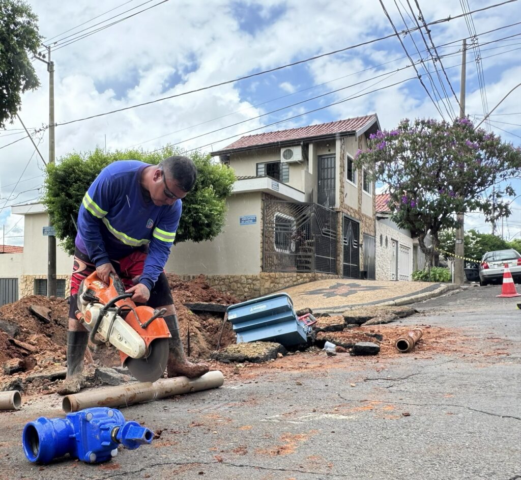 Um trabalhador da empresa de saneamento utiliza uma serra de disco motorizada para cortar uma tubagem de PVC em pleno pavimento da rua. Em primeiro plano, no chão, sobressai uma válvula/registro hidráulico de cor azul.
