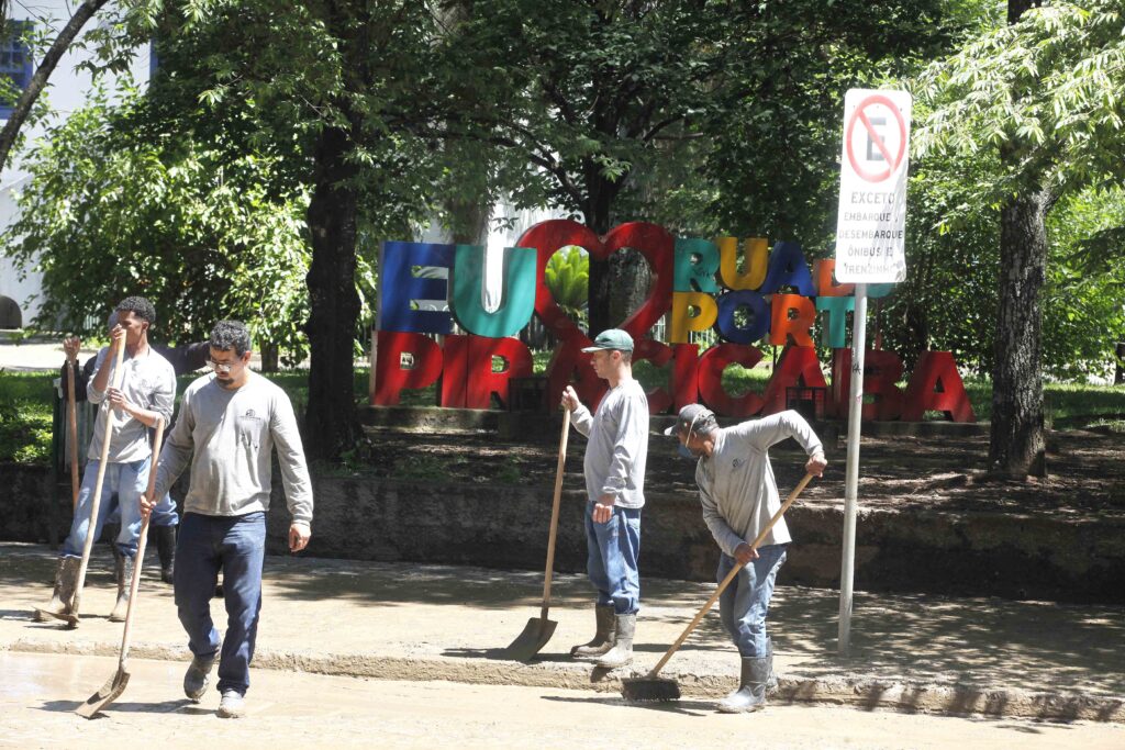Quatro trabalhadores utilizam pás e rodos para raspar a lama acumulada numa via pública, tendo ao fundo o letreiro colorido "EU AMO RUA DO PORTO PIRACICABA" e uma placa de trânsito.