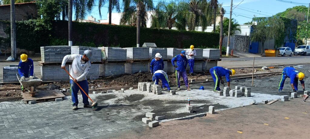 Vários trabalhadores com fardamento azul e capacetes de proteção a instalar blocos intertravados de betão e a varrer a área em obras.