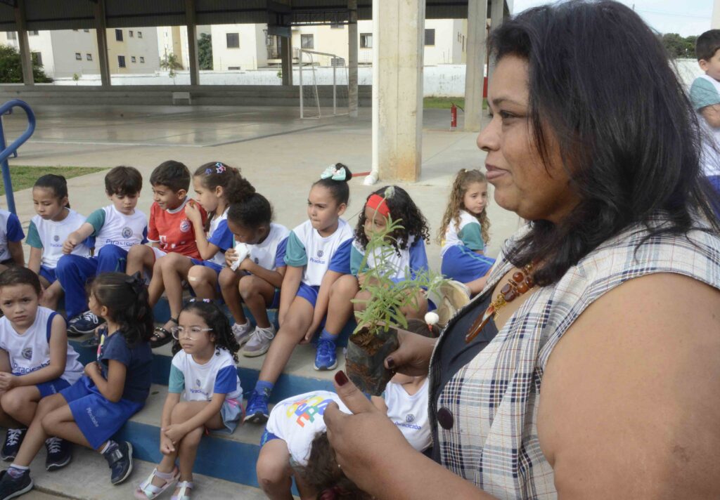 A educadora Sirlene Santos segura um vaso com uma muda de planta em primeiro plano enquanto fala com um grupo de alunos sentados nas bancadas de uma área coberta ao fundo.