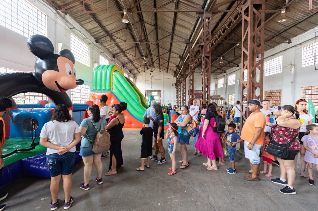 A imagem mostra um evento recreativo infantil realizado em um amplo galpão industrial, com estrutura metálica aparente, pé-direito elevado e grandes janelas que permitem a entrada de luz natural. No espaço, dezenas de pessoas, entre adultos e crianças, formam filas para utilizar brinquedos infláveis coloridos, entre eles um grande castelo inflável com a figura de um personagem de desenho animado e um escorregador inflável de grande porte nas cores verde, amarelo e laranja. O ambiente apresenta intensa circulação de visitantes, sugerindo uma atividade comunitária voltada ao lazer e à convivência familiar, com diversas crianças aguardando sua vez de brincar enquanto os responsáveis acompanham a movimentação.